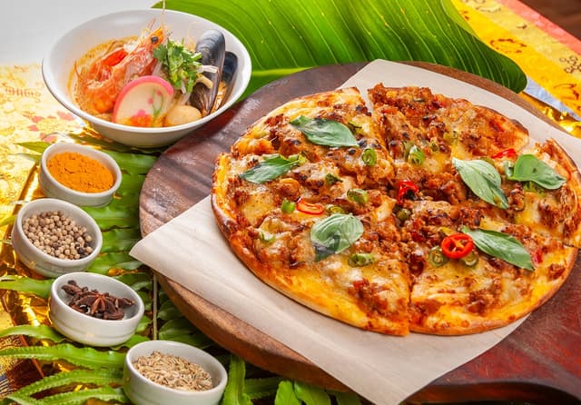 現煮「南洋叻沙麵」（左）湯頭辛香，入口微帶椰香甜味；「打拋豬肉披薩」（右）將經典熱炒搬上餅皮，是令人驚喜的東西合璧創意之作。／台北凱撒大飯店提供